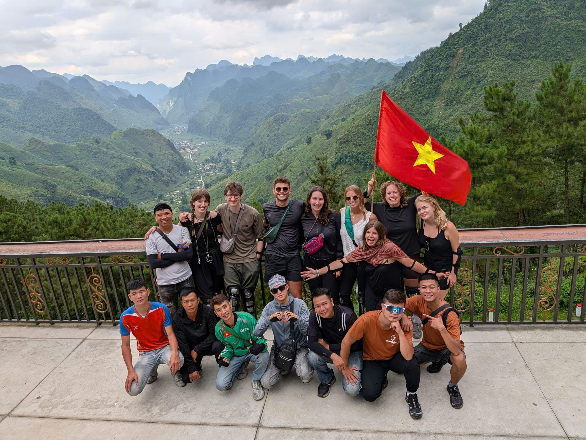 Travelers pose for photos with their local drivers at a scenic viewpoint overlooking a vast green valley surrounded by towering mountains.
